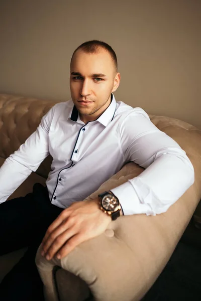 Husband at the photo session in studio. Wedding married groom. Man sitting at the sofa, dressed in white classic suit shirt with wrist watch looking at the camera, copy space.