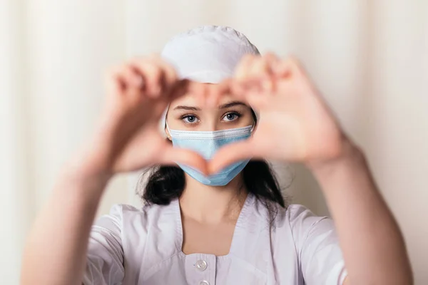 Nurse young medic standing show heart symbol shape by hands. Female doctor dressed in medical gown and surgical hat face blue mask. Image with copy space. Pandemic virus coronavirus concept