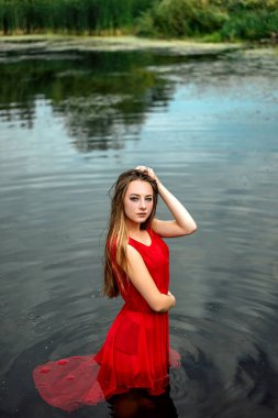 Su nehrinde ya da gölde dikilen, kırmızı elbiseli genç, ıslak kadın deniz kızının dikey portresi. Kameraya, fotokopi alanına ve doğanın arka planına bakıyor..