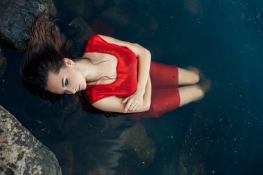 Su nehrinde ya da gölde kayaların üzerinde yatan, kırmızı elbiseli, yüzünü çevirmiş, fotokopi çeken ve tabiatın arka planını bulanık gösteren genç ve güzel bir kadın denizkızı portresi..