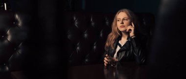 Bir bardak viskiyle yalnız oturan genç bir kızın geniş açılı görüntüsü. Deri ceketli kadın seçici odağı. Kopyalama alanı olan resim.