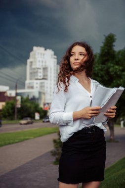 Genç, güzel, kıvırcık bir kadının dikey portresi. Elinde belgelerle dışarıda duruyor ve başka tarafa bakıyor, gülümsüyor. Beyaz tişörtlü ve siyah etekli bayan iş ortağını iş yerinde bekliyor..
