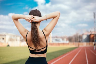 Arkadan bakan genç atletik kız at kuyruğu örüyor, vücudunu verimli bir spor koşusu için hazırlıyor. Şehir stadyumunda bayan sporcu modeli. Kopyalama alanı olan resim.