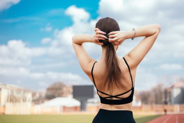 Arkadan bakan genç atletik kız at kuyruğu örüyor, vücudunu verimli bir spor koşusu için hazırlıyor. Şehir stadyumunda bayan sporcu modeli. Kopyalama alanı olan resim.