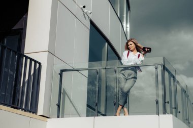 Ofis binasının yanında duran, el çantası taşıyan, güneş gözlüklü, beyaz gömlek ve pantolon giymiş, gözlerini kaçıran genç bir kadının tam boy portresi. Kopyalama alanı olan resim.