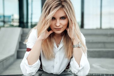 Beton merdivenlerde oturan kadın kameraya bakıyor. Beyaz şık gömlekli bayan koronavirüs salgını nedeniyle karantina sırasında dışarıda vakit geçiriyor. Küçük işletme moda kavramı. 