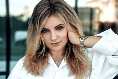 Dışarıda dikilen şık, güzel bir kadının yakın portresi. Kameraya bak. Beyaz parlak tişörtlü bayan, elinde gümüş saatle. Ofise yakın küçük bir işletme sahibi. Moda konsepti.