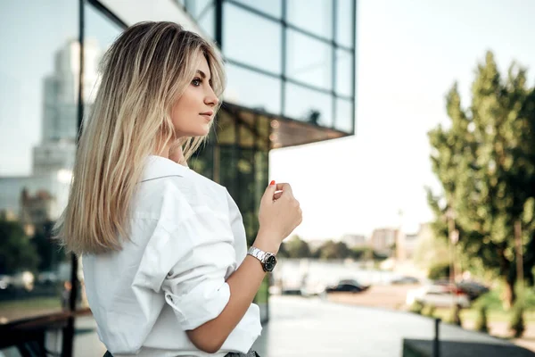 Modaya uygun güzel bir kadın dışarıda kameraya bakıyor. Elinde beyaz, parlak, gümüş bir saat olan bir bayan. Ofis binasının yakınında küçük bir işletme sahibi. Moda konsepti