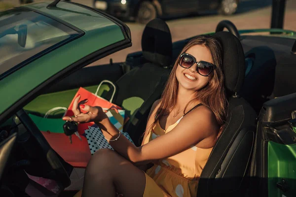 Close up portrait of woman sitting in the cabriolet car with shopping bags on the passenger seat, holding keys and smiling. Black friday concept.