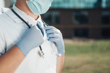 Hastanenin yanında steteskopla duran doktorun seçici odak noktası. Adam viral mavi maske, beyaz önlük ve eldiven takıyor. Sağlık ekibi dışarıda, anlaşıldı. Coronavirus konsepti.