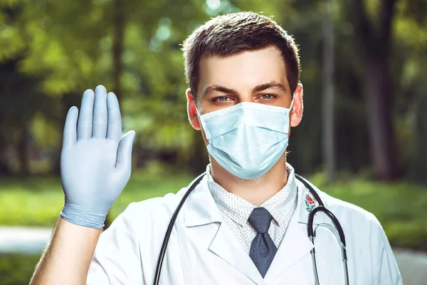 外に立っている医師が腕を上げてカメラを見ている 男は制服 青い顔のマスク 聴診器と手袋を医療ガウンを着用してください 健康保護の概念 コピースペース付き画像 — ストック写真