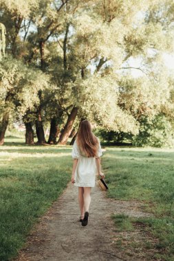 Uzun saçlı, beyaz elbiseli bir kadının dikey arka portresi. Yaz ormanlarında, parkta ya da çimlerin ya da çimenlerin yanındaki ormanda yürüyor. Hasır şapka takıyor..