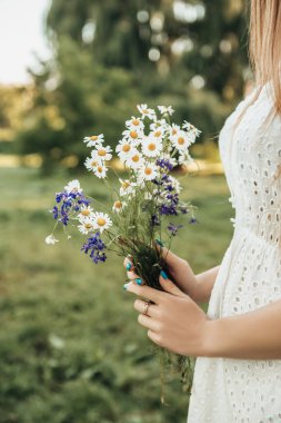 Yaz kenti parkında ya da orman ormanlarında yalnız başına duran yalnız kadın kır çiçeklerini tutuyor. Beyaz elbiseli bir bayan. Romantik bir kavram. Bulanık arkaplanı olan resim.