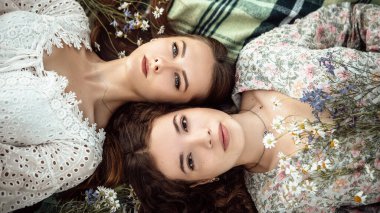 Ekosenin üzerine uzanmış renkli kır çiçekleriyle kameraya bakan iki güzel bayanın üst manzarasını kapatın. Yaz elbisesi giymiş kızlar. Romantik konsept. 