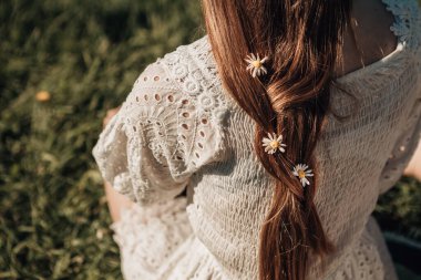 Güneşli bir yaz sabahında çimlerin üzerinde oturan genç bir bayanın nehir ya da göle bakışı. Parlak elbiseli, örgülü saçlı, papatyalı, doğa geçmişi olan bir kız..