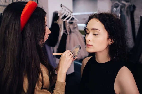 Make up talanted artist doing maquillage to woman applying concealer. Visagist using brushes shadows facial professional cosmetics for makeup. Beauty industry studio, image with copy space.