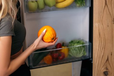 Güzel sarışın kadın açık buzdolabına meyve ve sebzelerle bakıyor. Eğitimden sonra kahvaltıya hazırlanan bayan, sağlıklı yemekler pişirmek istiyor..