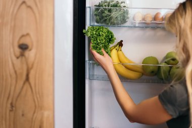 Güzel sarışın kadın açık buzdolabına meyve ve sebzelerle bakıyor. Eğitimden sonra kahvaltıya hazırlanan bayan, sağlıklı yemekler pişirmek istiyor..