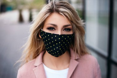 Çekici sarı saçlı bayan, tıbbi moda maskesi takıyor. Pembe, ılık, sonbahar bahar montu giyiyor. Kamera koronavirüs karantina konseptine bakıyor.