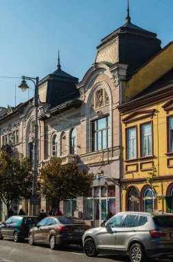 Targu Mures, Romanya - Ekim 17.2025. Romanya 'nın Targu Mures kentinin tarihi merkezinde, parlak mavi gökyüzü altında süslü binalar, sokak parkı, trafik ve sonbahar ağaçlarının yer aldığı bir sokak manzarası