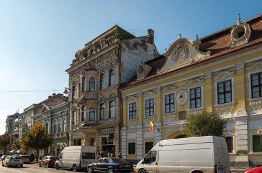 Targu Mures, Romanya - Ekim 17.2025. Romanya 'nın Targu Mures kentinin tarihi merkezinde, parlak mavi gökyüzü altında süslü binalar, sokak parkı, trafik ve sonbahar ağaçlarının yer aldığı bir sokak manzarası