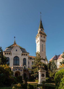 Targu Mures, Romanya - Ekim 17.2025. İkonik Kültür Sarayı ve Romanya 'nın Targu Mures kentinin Eski Belediye Kulesi, işlek bir cadde boyunca parlak mavi bir gökyüzünün altında ağaçların altında duruyor.