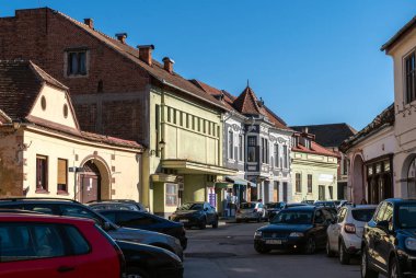 Medya, Romanya - Ekim 20.2025. Romanya 'nın Medias kentinde klasik Sakson mimarisi, kırmızı kiremitli çatılar ve park halindeki arabalarla renkli tarihi cephelerin yer aldığı bir caddenin güneşli perspektifi