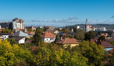 Alba Iulia, Romanya - Ekim 19.2025. Romanya 'da mavi bir gökyüzü altında sonbahar ağaçları tarafından çerçevelenmiş kırmızı kiremitli çatılar ve çeşitli mimarileri gösteren manzaralı bir kent manzarası