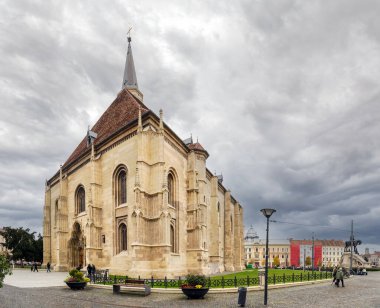 Cluj-Napoca, Romanya - Ekim 18.2025. Gotik St. Michael Kilisesi ve merkez Union Square of Cluj-Napoca 'daki Matthias Corvinus Heykel Grubu' nun gri gökyüzü altında geniş bir manzarası vardır.