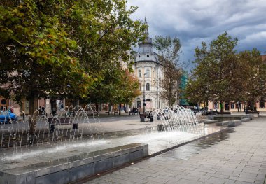 Cluj-Napoca, Romanya - Ekim 18.2025. Modern bir yer çeşmesi tarihi binalar ve sonbahar ağaçlarıyla çevrili bir yaya meydanına su püskürtür.