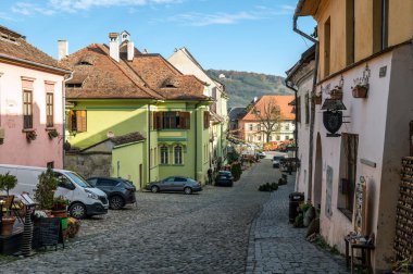 Sighisoara, Romanya - 21.10.2025. UNESCO 'nun ortaçağ Sighisoara kalesinde dar bir kaldırım taşı geçidi boyunca uzanan parlak pembe, yeşil ve turuncu tarihi evlerin büyüleyici manzarası.