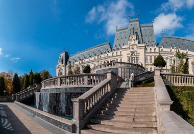Iasi, Romanya - 23 Ekim 2025. Iasi Romanya 'daki Neo-Gotik Kültür Sarayı' nın geniş panoramik manzarasında, parlak mavi gökyüzü altında fıskiyeler ve sonbahar ağaçları bulunan bereketli Palas Halk Bahçesi yer alıyor.