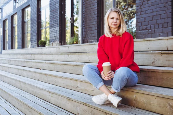 Kadın merdivenlerde oturuyor ve elinde tek kullanımlık bir fincan kahve tutuyor..