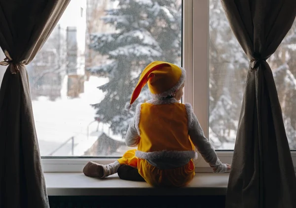 小さな男の子は窓の外に座って黄色の服を着て冬の雪が降る景色を見て 家で幸せな家族の時間 — ストック写真