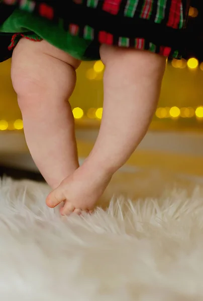 女婴穿着她的第一套红绿相间的天鹅绒裙 光溜溜的腿和脚趾头在温暖的花环背景下触摸毛毯 — 图库照片