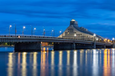 Aydınlatılmış Taş Köprü (Akmens eğikliği) ve modern Letonya Ulusal Kütüphanesi (Gaismas pils) binası, alacakaranlıkta, derin mavi bir gökyüzünün altında Daugava Nehri 'ne yansıtılır..