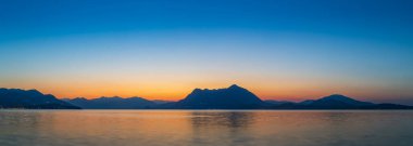 Maggiore Gölü 'nün panoramik manzarası ve Alpler ve dağların silueti (muhtemelen Mottarone), Stresa' dan gelen parlak turuncu ve mavi renkleri yansıtır.