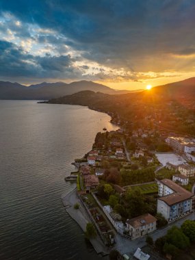 Güneş doğarken Maggiore Gölü 'ndeki Porto Valtravaglia kıyı kasabasının havadan görünüşü limanı, renkli binaları ve kasabanın arkasındaki sarp orman tepelerini gösteriyor.