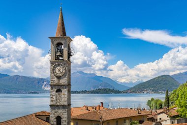 Laveno Mombello 'nun çatılarına bakın, uzun taş çan kulesi ve Sant' Ambrogio Kilisesi cephesi, Maggiore Gölü 'nün mavi suları ve bulutlu bir gökyüzünün altındaki uzak dağlar