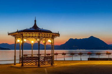 Morning view from the promenade featuring an illuminated ornamental cast-iron gazebo and Mount Mottarone in silhouette across Lake Maggiore, before a colorful twilight sky