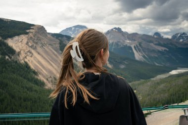 Ağaçlar ve bulutlu gökyüzü ile görkemli dağ manzarasına hayran bir kadın. Columbia Buz Sahası, AB
