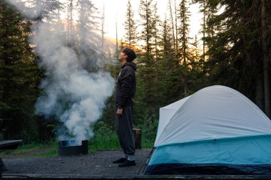 Bir kampçı kamp çadırının yanındaki ateşten yükselen dumana bakıyor. Takakkaw Şelalesi, BC