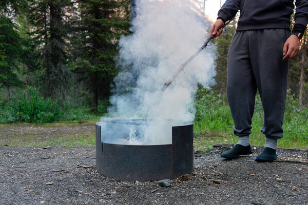 Person tending a smokey fire in a metal firepit in a forest area. Takakkaw Falls, BC