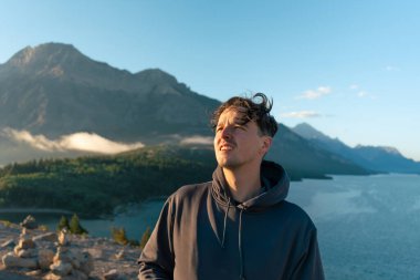 Kapüşonlu bir adam güneşli bir günde manzaralı dağ gölü manzarasına bakıyor. Waterton, AB