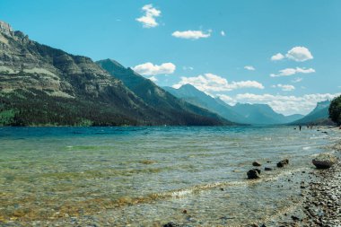 Rocky kıyı şeridi, heybetli dağların altında sakin gölle buluşur. Waterton, AB