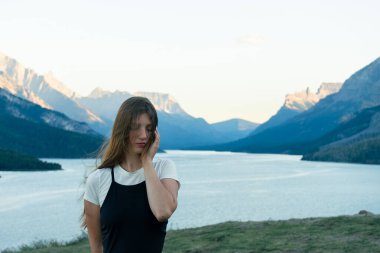 Huzurlu bir kadın görkemli dağların ve parıldayan bir gölün önünde duruyor. Waterton, AB