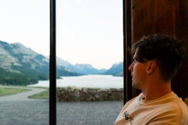 Bir adam pencereden göle bakıyor. Etrafı resimli dağlarla çevrili. Waterton, AB