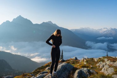 Aktif kıyafetli bir kadın manzaralı bir dağın tepesinde duruyor. Buzul Ulusal Parkı, BC