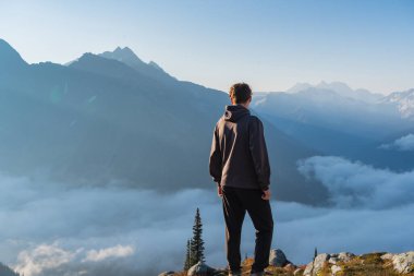 Bir dağ zirvesinde, ilerideki sisli dağlara bakan bir adam. Buzul Ulusal Parkı, BC