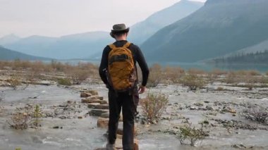 Bir yürüyüşçünün sığ bir derenin üzerinde yol oluşturan yerleştirilmiş kaya parçalarının gerçek zamanlı videosu. Berg Gölü ve Alp manzarası Robson İl Parkı 'ndan görülebiliyor..
